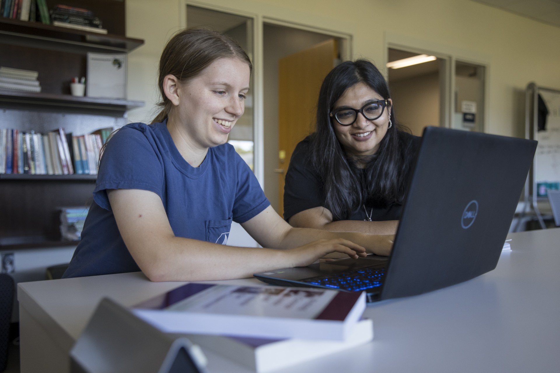 Two people sit at a table using a laptop, with one person typing on the keyboard while the other looks at the screen. Books are stacked on the table, and shelves and office doors are visible in the background.